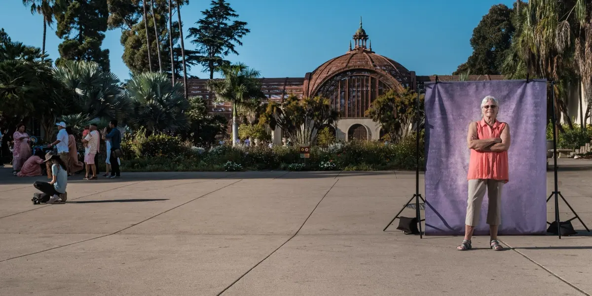 Cheryl crosses her arms with a steady smile, in sunglasses and a coral sleeveless top, standing at a purple backdrop on the Balboa Park promenade near the Botanical Building.