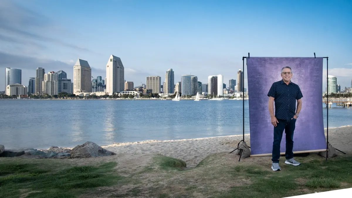 Chris stands relaxed in a navy dotted shirt at a purple backdrop on the shoreline, with a sweeping view of San Diego Bay and the downtown skyline beside him.