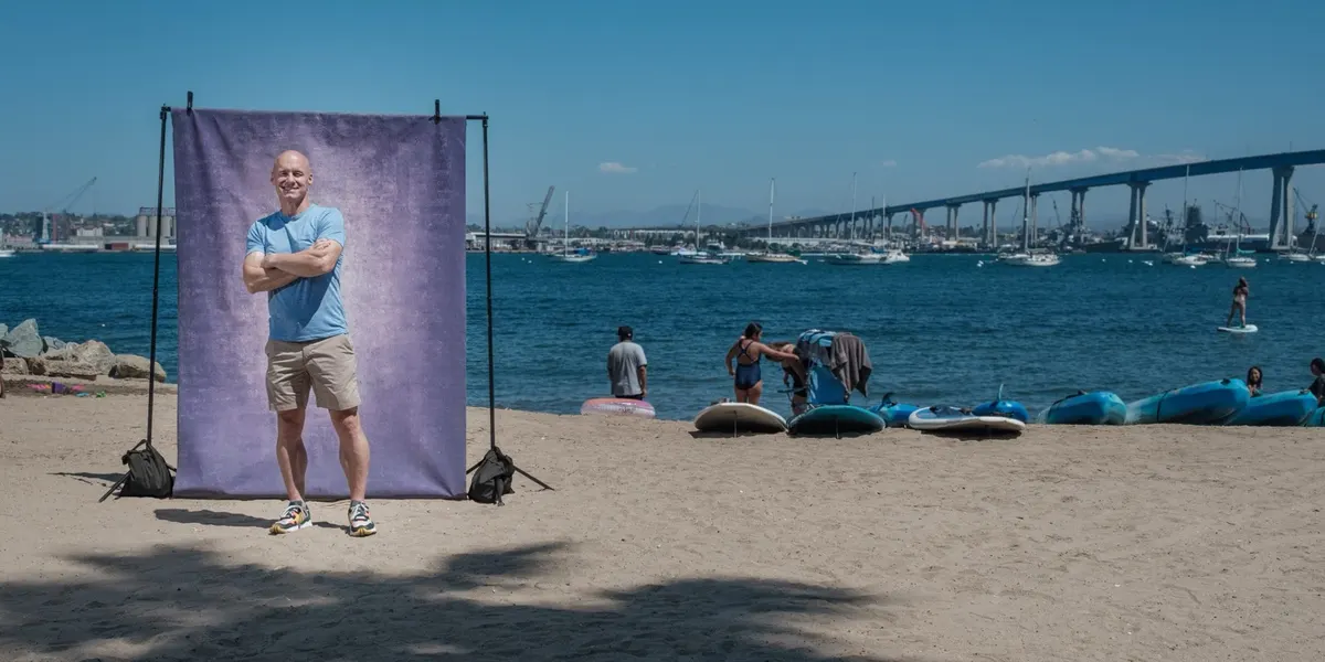 CJ grins with his arms crossed in a light blue tee, standing in front of a purple backdrop on the beach with kayaks and the Coronado Bridge behind him.
