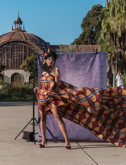 Tootie poses in a sculpted headpiece as a sweeping fan-print gown billows out beside her, in front of a purple backdrop near the Balboa Park Botanical Building.