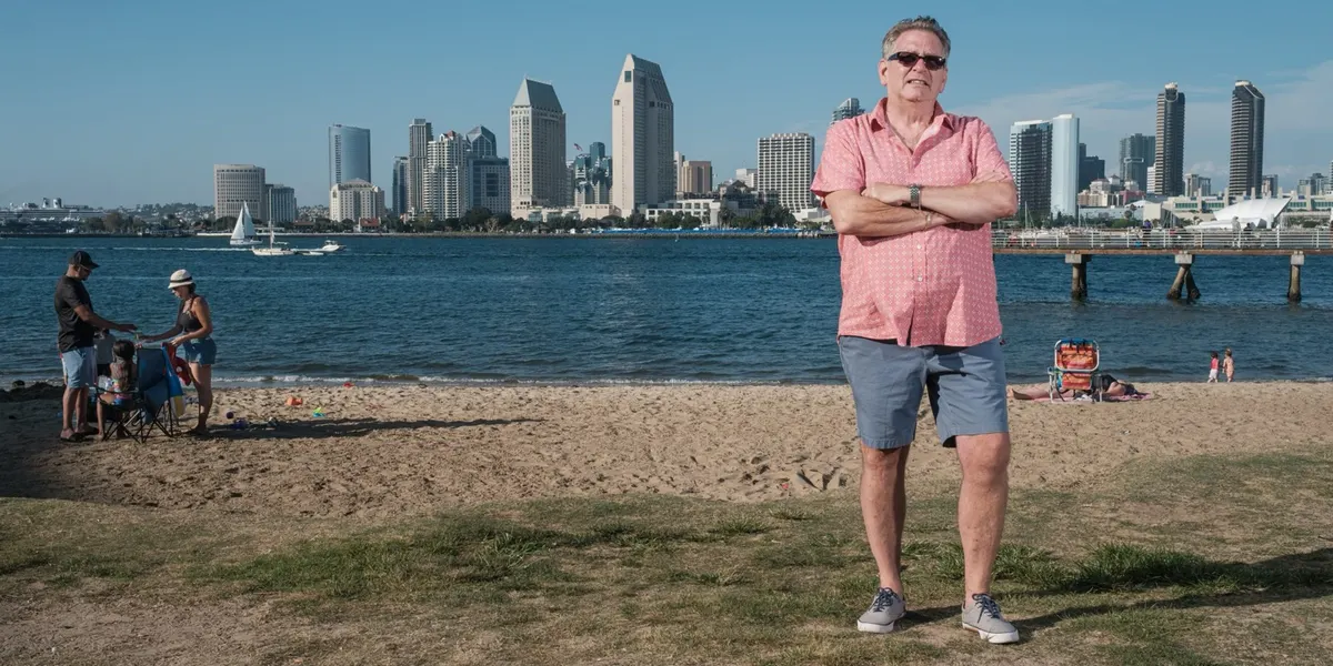 Jeff stands with arms crossed in sunglasses and a coral patterned shirt on a grassy rise above the beach, the San Diego skyline shining across the bay behind him.