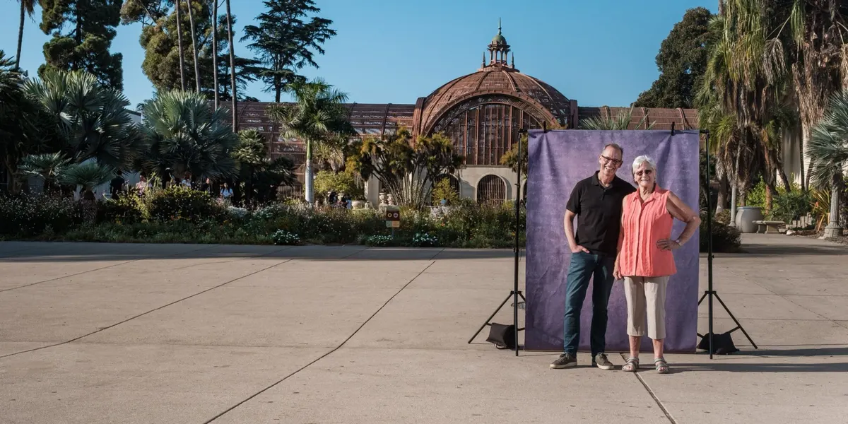 Michael and Cheryl stand arm in arm and smiling, he in a black polo and she in a coral top, in front of a purple backdrop at the Balboa Park Botanical Building.