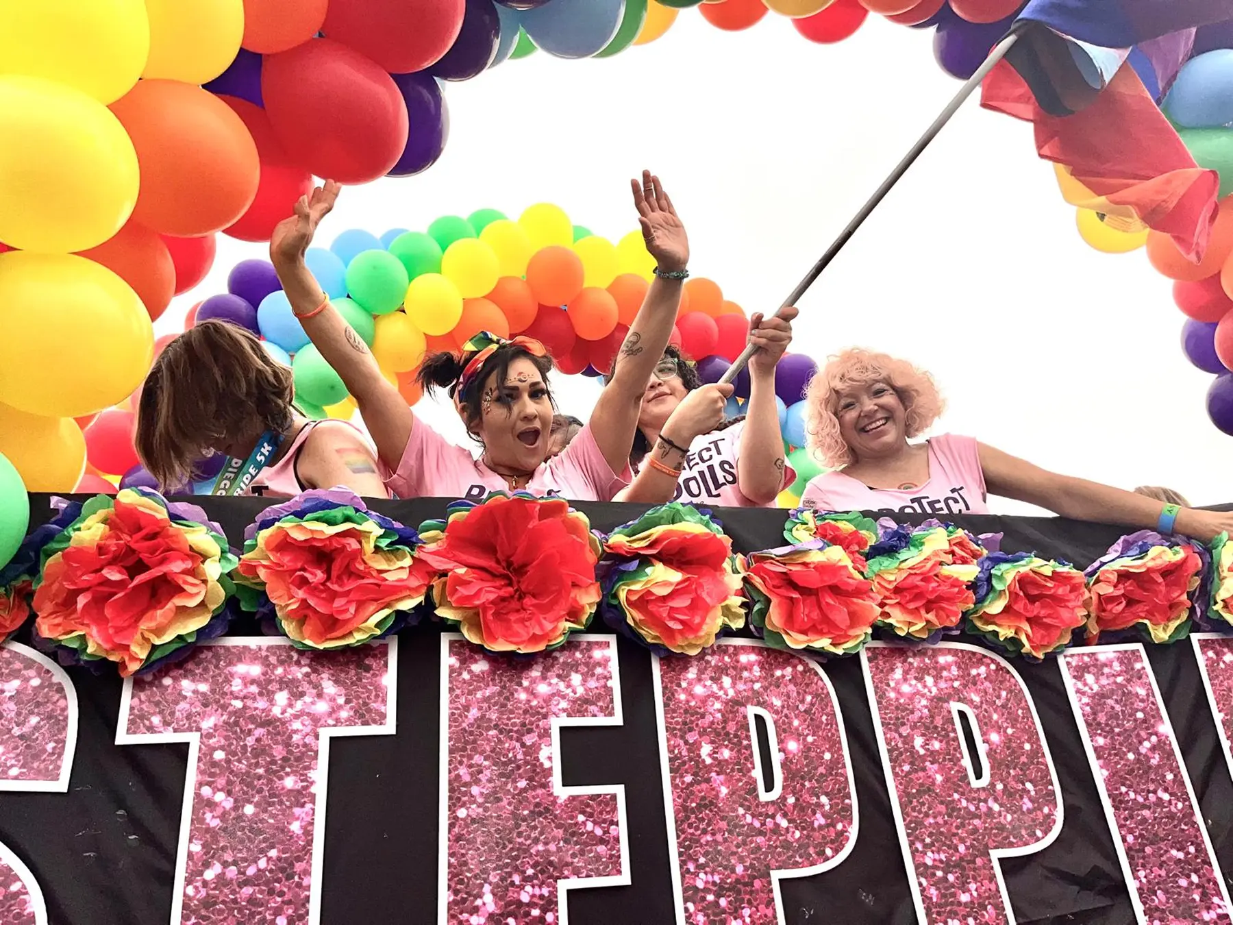 Stepping Stone team waving from their Pride float beneath a rainbow balloon arch