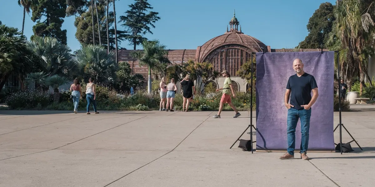 Tim stands easy with thumbs in his jean pockets, wearing a navy tee, in front of a purple backdrop on the Balboa Park promenade with the Botanical Building dome rising behind him.