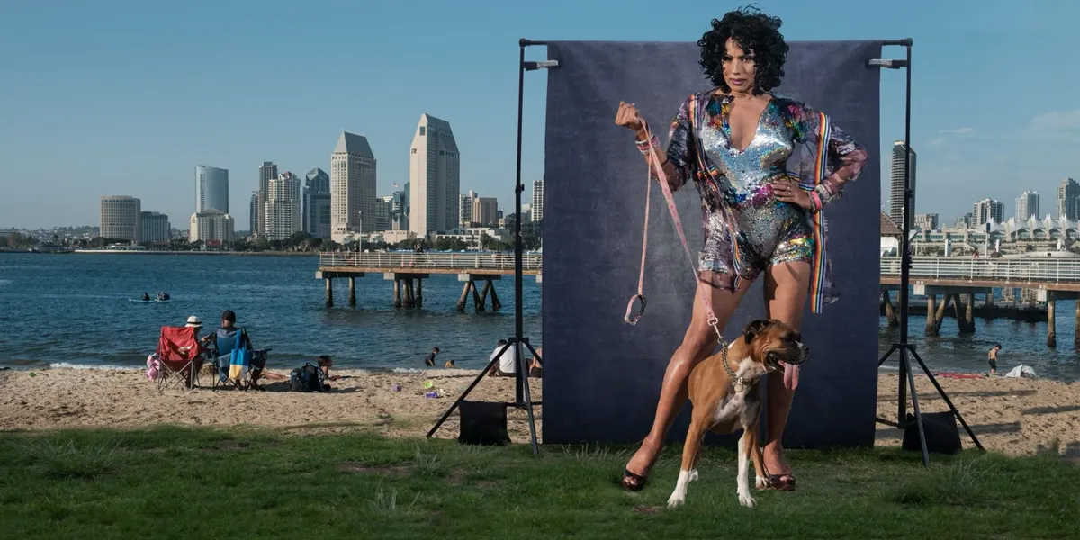 Tootie sparkles in a sequined romper with a rainbow-trimmed wrap, holding the pink leash of a happy boxer dog, in front of a navy backdrop with the San Diego skyline across the water.